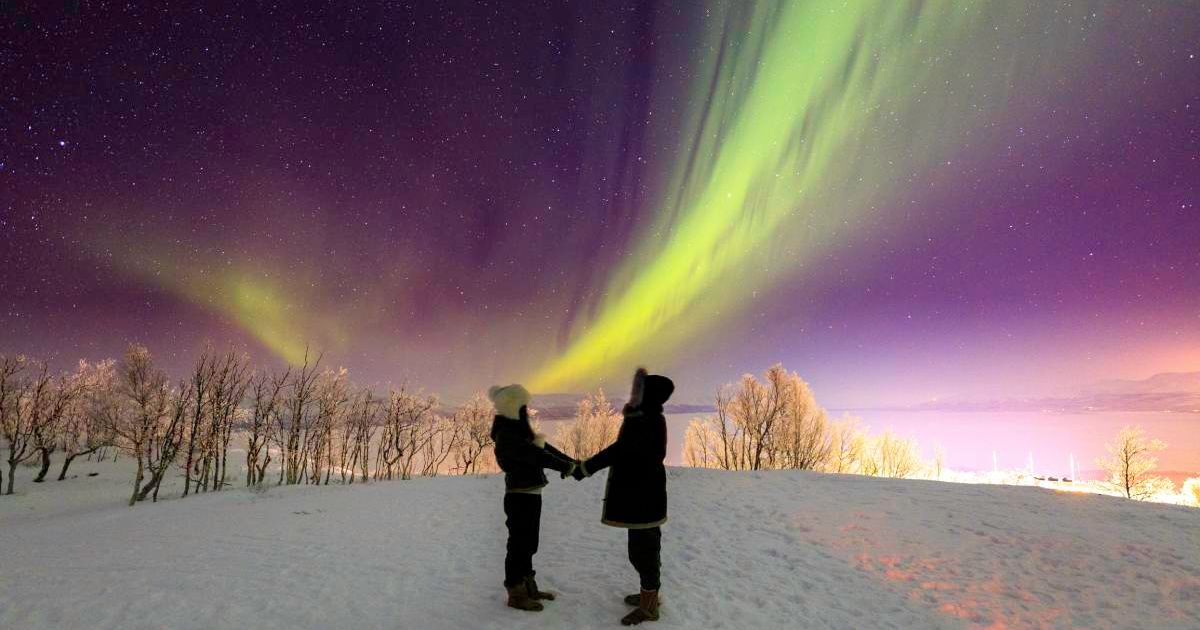 Two people standing hand in hand beneath a dazzling display of Northern Lights (Representative Cover Image Source: Getty Images | Samuli Valnionpaa)