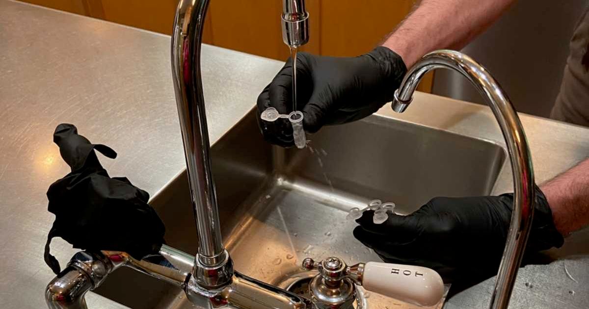 A USGS scientist wearing black gloves is collecting a sample of tap water from the kitchen sink using small plastic vials to test for PFAS. (Image Source: USGS)